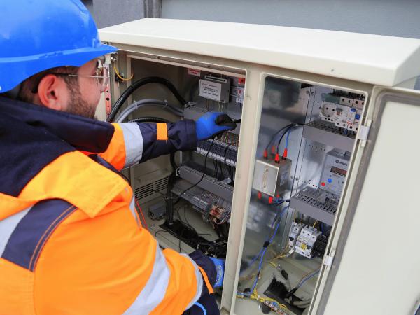 Elektryk w kasku i odzieży ochronnej wykonuje pomiary w szafie sterowniczej. Okresowy przegląd techniczny instalacji elektrycznej, rozdzielnicy z aparaturą modułową i licznikami.