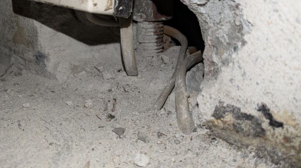 Zdjęcie przedstawia fragment starej instalacji elektrycznej wewnątrz surowej, betonowej wnęki. Przez otwory w betonie przechodzą płaskie przewody w jasnej izolacji, które są silnie zakurzone i pokryte gruzem. W głębi widoczna jest metalowa, gwintowana śruba lub pręt, a całe otoczenie sprawia wrażenie zaniedbanego i wymagającego remontu.