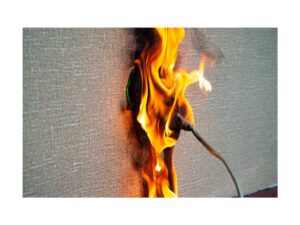 Zdjęcie pożaru gniazdka elektrycznego w ścianie pokrytej tapetą. Intensywne pomarańczowe płomienie wydobywają się z wnętrza puszki, obejmując podłączoną wtyczkę i wypalając pionowy pas w materiale tapety