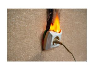 Białe gniazdko elektryczne na ścianie z tapetą, z którego wydobywają się płomienie. Górna część obudowy gniazdka jest stopiona, a nad nim widoczny jest pionowy, czarny ślad osmalenia na tapecie. Do dolnego wejścia podłączona jest wtyczka z kablem.