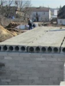 Zdjęcie przedstawia plac budowy, na którym widoczne są prefabrykowane płyty kanałowe ułożone na murowanej ścianie budynku. Płyty mają charakterystyczne podłużne otwory (kanały) widoczne na ich przekroju czołowym. W tle widać pracującego mężczyznę, nasyp ziemi oraz inne zabudowania mieszkalne.